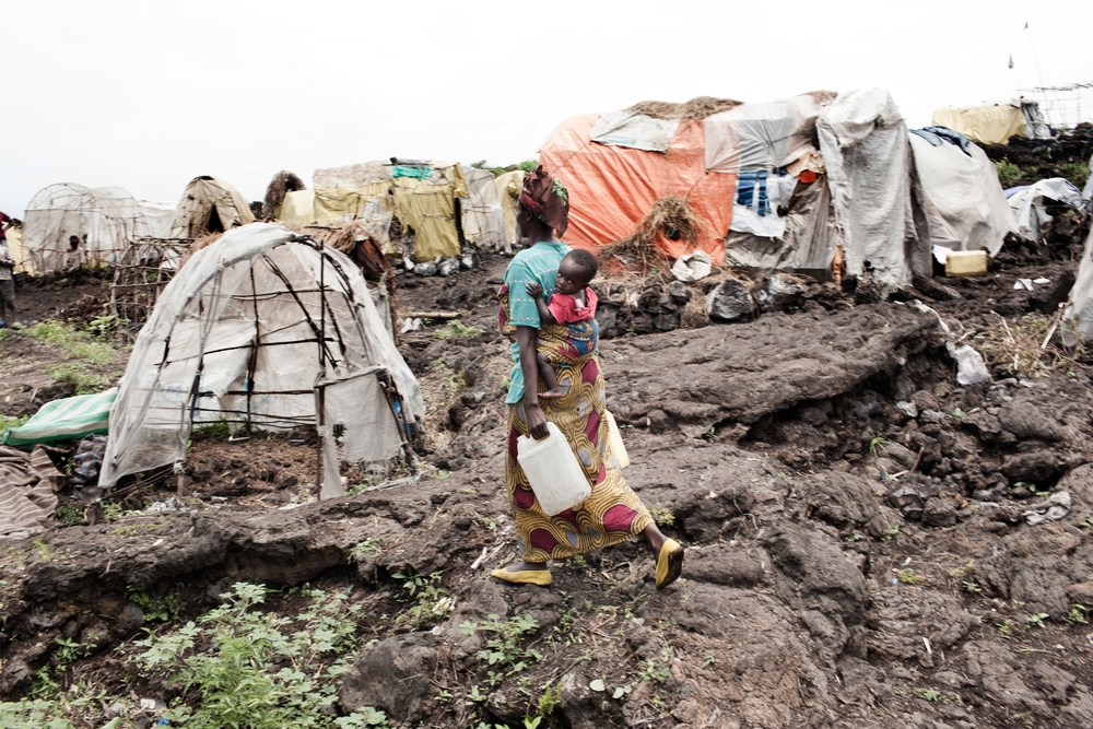 A woman carrying a baby on her back and two water containers walks across rocky ground towards makeshift tents.