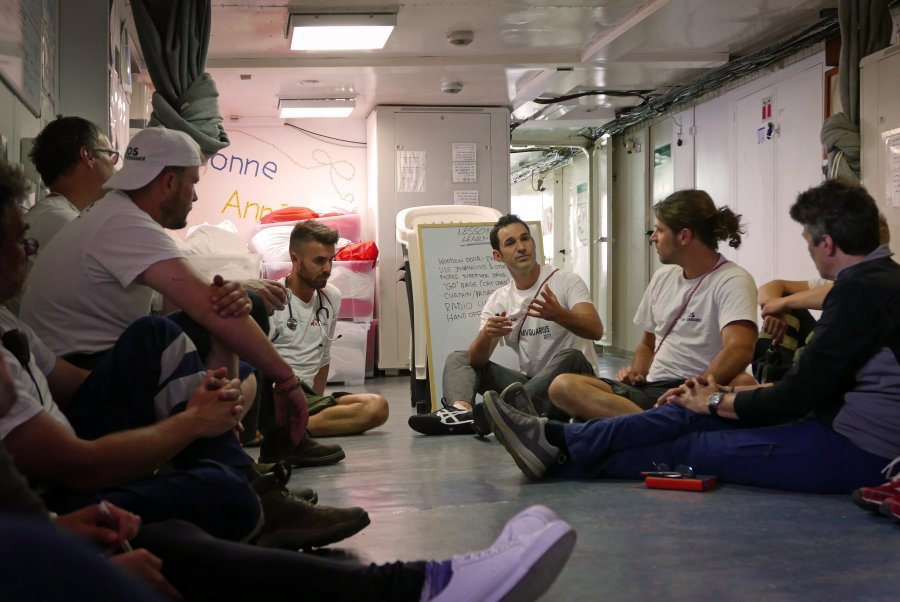 While the team wait for a rescue out in international waters, in the women's shelter, MSF doctors Conor Kenny and Craig Spencer carry out training for the non-medics on board in how to assist in case of a cervical spine injury.