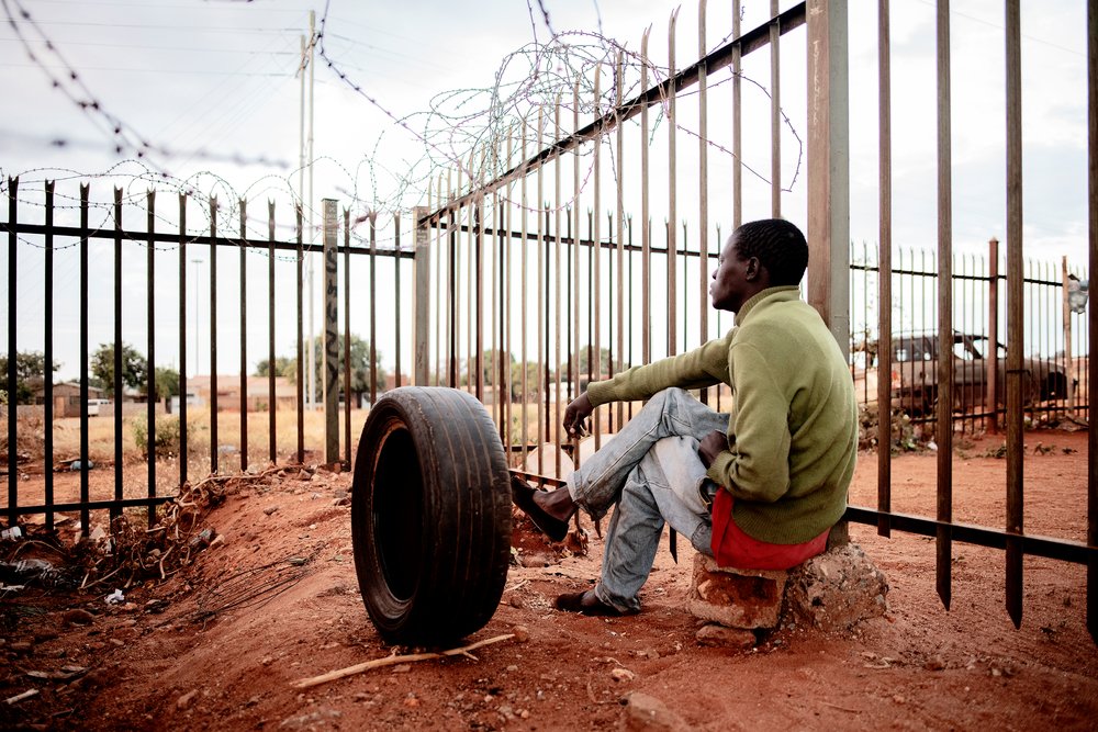 A man is seen looking through the fence line at a temporary shelter where migrants entering South Africa through Musina take refuge. © Luca Sola 2019
