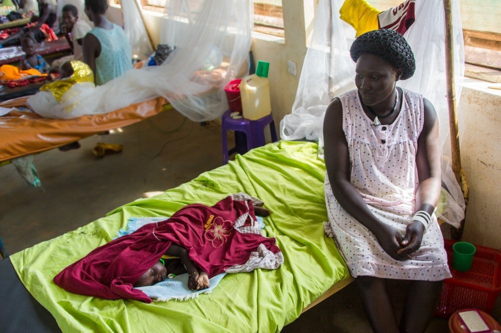 A 1-year-old baby sleeps at MSF&#039;s Therapeutic Feeding Centre at Aweil while his mother looks on. MSF runs the 250-bed Aweil hospital, in Northern Bahr El Ghazl state, focusing on maternal healthcare and paediatrics, including nutrition.