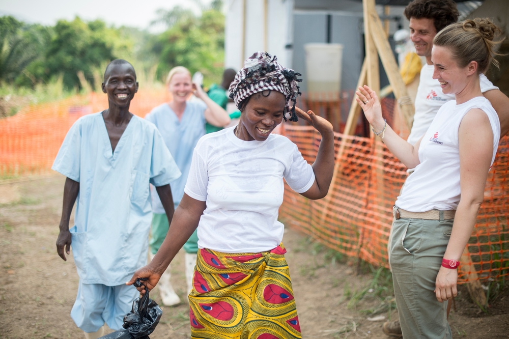 Two health workers in MSF shirts and two others in scrubs smile and wave at a woman.