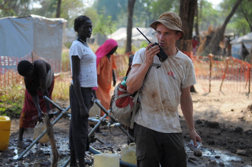 A man in an MSF shirt uses a 2 way radio, while in the background women fill containers with water