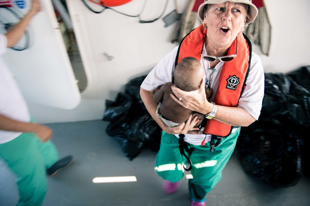 An MSF worker on a ship wearing a life jacket holds a small baby.