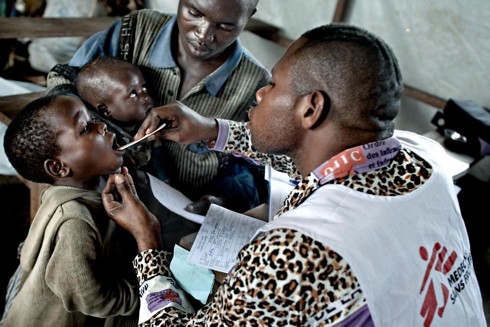 An MSF worker looks into the mouth of a child. A man in the background holds a baby.