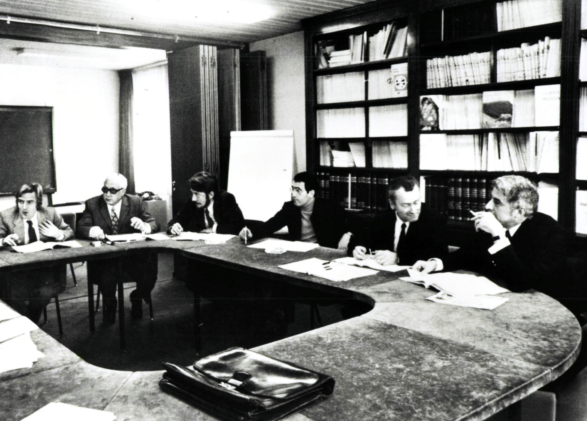 An old black and white photo of a group of men sitting around a large office table with bookshelves in the background.