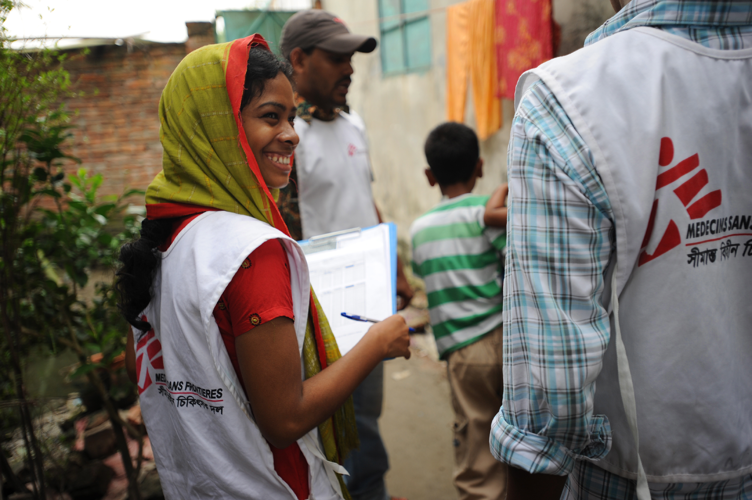 A woman wearing an MSF shirt and a head scarf holds a clip board and smiles at another MSF worker partially out of frame.